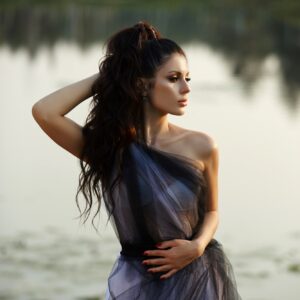 Woman in a long dress near the lake sits on an old boat, beautiful woman in rays of the evening sun